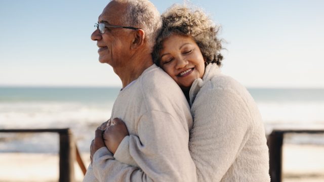 Affectionate senior woman smiling happily while embracing her husband by the ocean. Romantic elderly couple enjoying spending some quality time together after retirement.