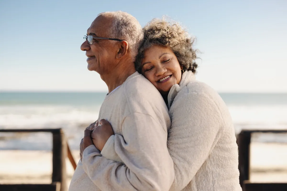 Affectionate senior woman smiling happily while embracing her husband by the ocean. 
