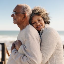 Affectionate senior woman smiling happily while embracing her husband by the ocean. Romantic elderly couple enjoying spending some quality time together after retirement.