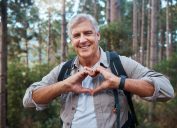 Portrait, heart and senior man in forest hiking, fitness and health support, self care hands and cardiovascular workout. Nature, travel or trekking of happy person in woods with love sign or emoji