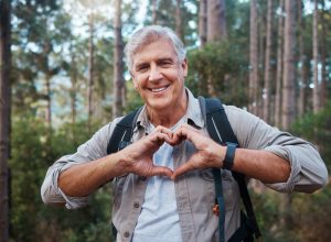 Portrait, heart and senior man in forest hiking, fitness and health support, self care hands and cardiovascular workout. Nature, travel or trekking of happy person in woods with love sign or emoji