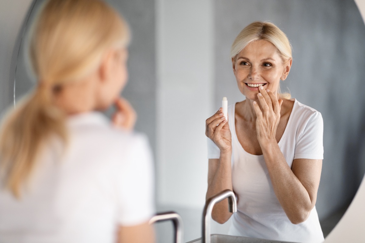 Beautiful Mature Woman Looking At Mirror And Applying Moisturising Lip Balm, Attractive Happy Senior Lady Holding Chapstick, Using Hygienical Lipstick While Making Domestic Beauty Routine