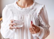 Woman in white blouse holding in hand iron Ferrum supplement capsule and glass of water. Bioactive additive woman pharmacy. Vitamin mineral treatment against anemia. Autumn health care concept