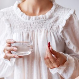 Woman in white blouse holding in hand iron Ferrum supplement capsule and glass of water. Bioactive additive woman pharmacy. Vitamin mineral treatment against anemia. Autumn health care concept