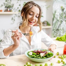 Young woman eating healthy food sitting in the beautiful interior with green flowers on the background