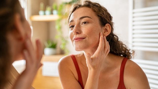 Beauty girl looking at mirror while touching her face and checking pimple, wrinkles and bags under the eyes, during morning beauty routine. Happy smiling beautiful young woman applying moisturizer.
