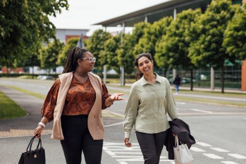 A three quarter length shot of two businesswomen walking and talking to each other after work. They are both dressed smartly and casually and are walking over a road. They are based in the North East of England.