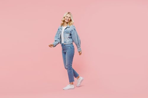 A happy middle-aged blonde woman wearing a denim jacket and jeans against a pink background.