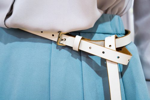 Close up of a woman's midsection wearing a light gray blouse, light blue trousers, and a cream-colored skinny belt