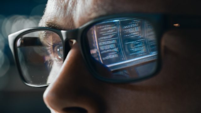 Close-up shot of a man wearing glasses where you can see the reflection of computer data in his lenses