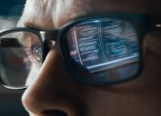 Close-up shot of a man wearing glasses where you can see the reflection of computer data in his lenses