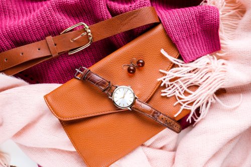 Close up of a bright pink sweater and light pink scarf with brown leather accessories including a belt, clutch, and watch