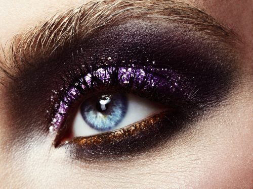 Close up of a woman's eye wearing heavy purple and gold glittery eye makeup with black liner
