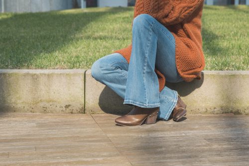 Close up of a woman sitting on a curb wearing a burnt-orange sweater, flare-leg jeans, and brown boots.