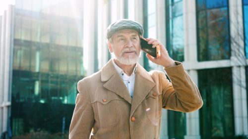 Portrait successful retired fashionable grey-haired man walking outside calling talking on smartphone. Stylish mature calm male wearing coat and cap speaking planning using technologies discussing.