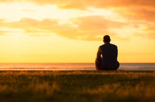 man sitting on a hill by himself looking out at the sunset