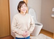 A content-looking young woman stands in her bathroom next to the toilet with her hands on her stomach