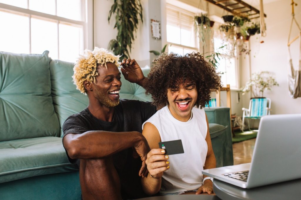 Two men sitting on floor in front of couch, shopping on laptop and laughing