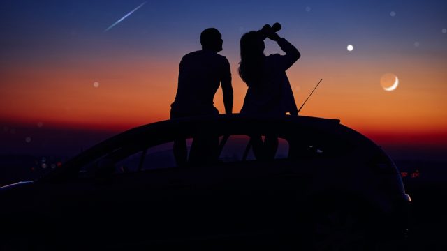 A couple sitting on top of a car stargazing with a pair of binoculars at dusk as a shooting star travels overhead