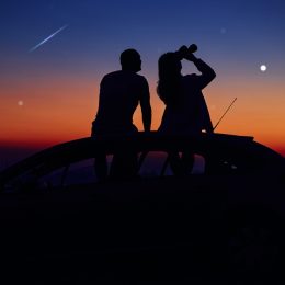 A couple sitting on top of a car stargazing with a pair of binoculars at dusk as a shooting star travels overhead