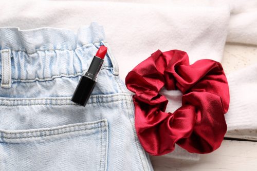Close up of elastic-waist light-wash jeans, a red scrunchie, and red lipstick against a white background