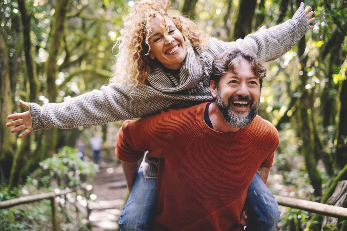 Overjoyed adult couple have fun together at outdoor park in leisure activity. Man carrying woman in piggyback and laugh a lot. Love and life mature people lifestyle concept. Enjoying vacation nature