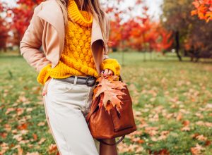 Close up of stylish woman holding handbag wearing yellow sweater in autumn park among red trees. Fall female clothes and accessories. Fashion. Space