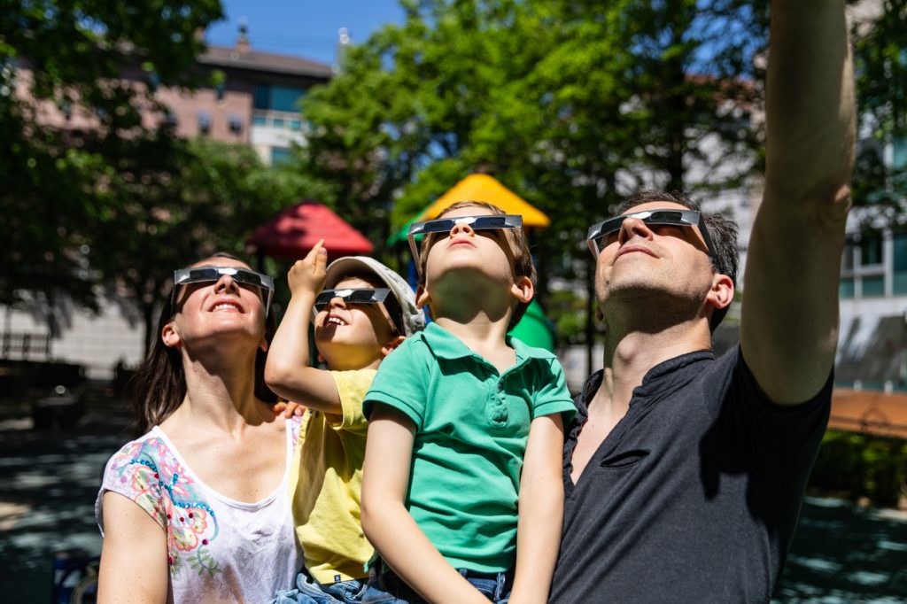 Parents holding two young children watching a solar eclipse while wearing protective glasses