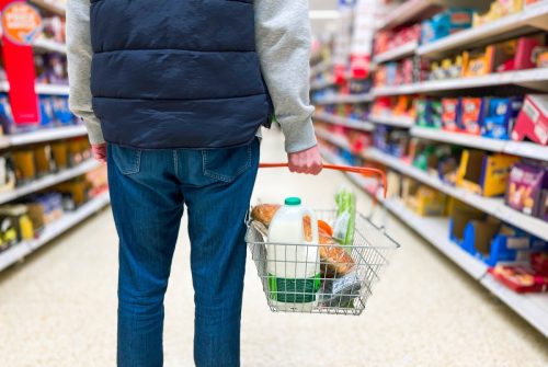 Low angle close up color image depicting a man holding a shopping basked filled with essential fresh groceries like bread and milk in the supermarket.
