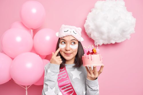woman holding a birthday cake against a pink backdrop with under eye masks on