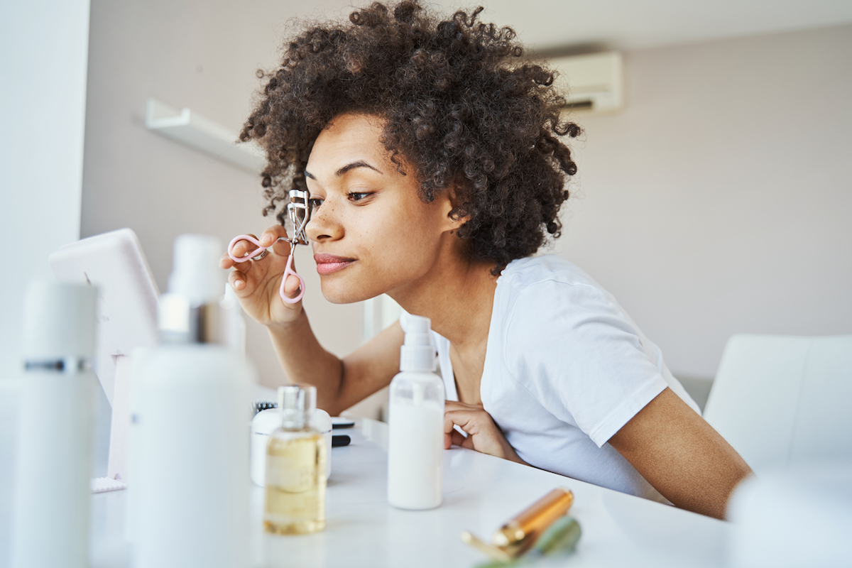 Tranquil concentrated African American woman curling her eyelashes with a cosmetic tool before the mirror