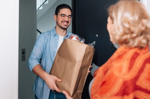 A young male volunteer handing a grocery bag at the door to an older woman
