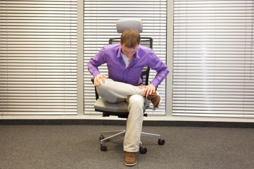 Man wearing khaki pant and a purple shirt sits in his office chair with his ankle crossed over his knee, stretching his hip
