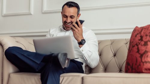 Portrait of a confident mature formal dressed man talking on mobile phone while sitting in the hotel room with laptop. Businessman on tour working from hotel room.
