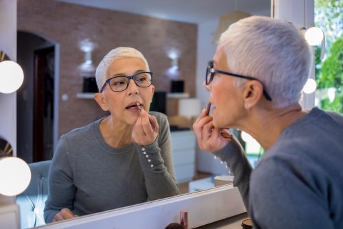 Portrait of smiling senior woman with short grey hair looking at herself in the mirror and putting lipstick on.