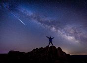 A person standing on top of a hill watching a shooting star from a meteor shower and looking at the Milky Way