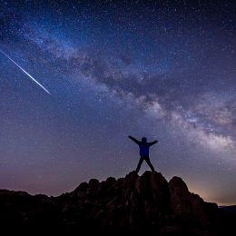 A person standing on top of a hill watching a shooting star from a meteor shower and looking at the Milky Way