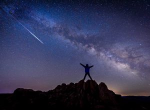 A person standing on top of a hill watching a shooting star from a meteor shower and looking at the Milky Way