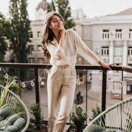 Stylish young woman on balcony in beige neutral outfit with blouse and high-waisted trousers