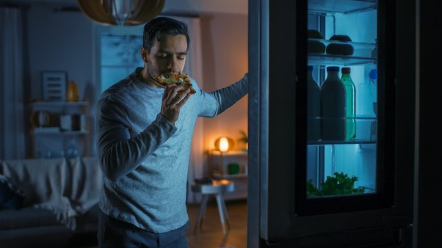 Sleepy man eating pizza next to open fridge at night