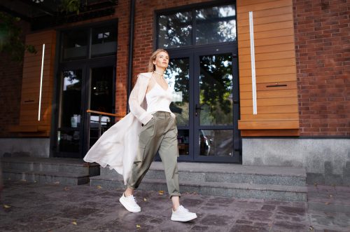 A young, stylish woman is walking down a city street wearing a white silk tank, sheer white long open shirt, green cargo pants, and white sneakers