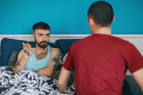 man in bed looking at his boyfriend and explaining, using hand gesture