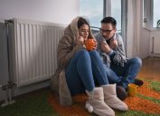 Young couple in jacket and covered with blanket sitting on floor beside radiator and trying to warm up