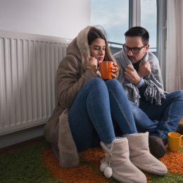 Young couple in jacket and covered with blanket sitting on floor beside radiator and trying to warm up