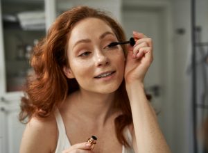 woman with red hair applying mascara in mirror