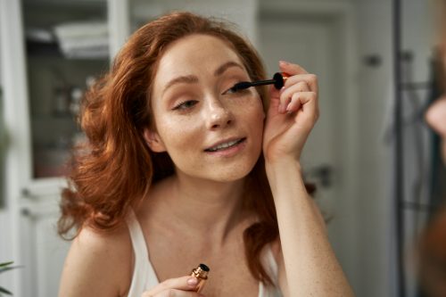 woman with red hair applying mascara in mirror