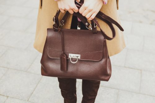 Female hands holding a brown leather handbag wearing a camel-colored coat