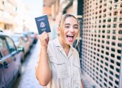 A young woman holding up a passport on the street while smiling