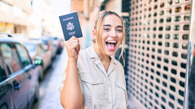 A young woman holding up a passport on the street while smiling
