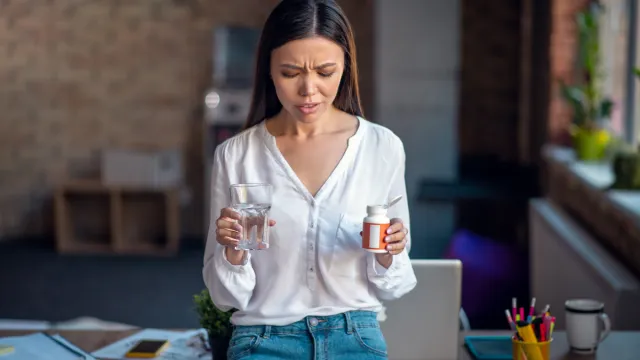 A young woman who's not feeling well leans against her desk with a glass of water in one hand and a pill bottle in the other